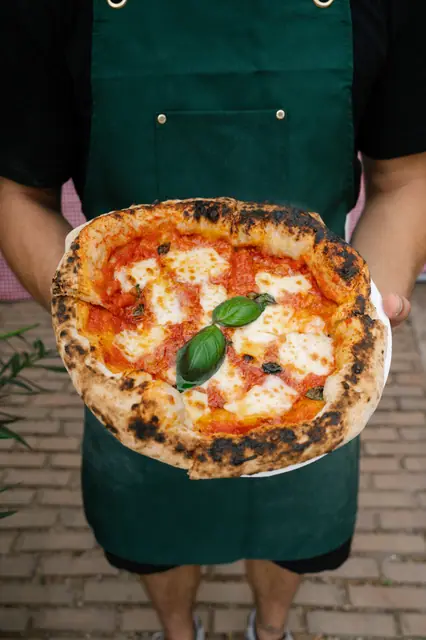Pizzaiolo v zelené zástěře drží Margheritu s bazalkou